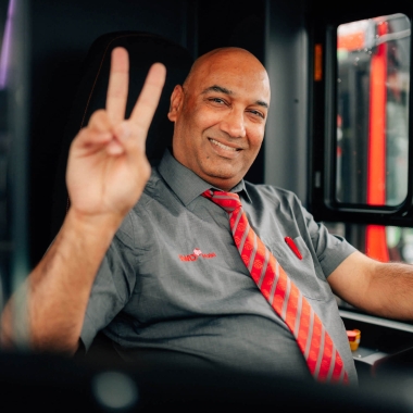 Ein Busfahrer von SWO Mobil im grauen Hemd mit rot-gestreifter Krawatte sitzt am Steuer eines Busses, lächelt und zeigt das Peace-Zeichen in die Kamera.