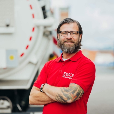 Porträt eines lächelnden Mitarbeiters der SWO Netz GmbH mit Bart und Brille im roten Poloshirt, der mit verschränkten Armen vor einem weißen Sonderfahrzeug steht. Querformat.