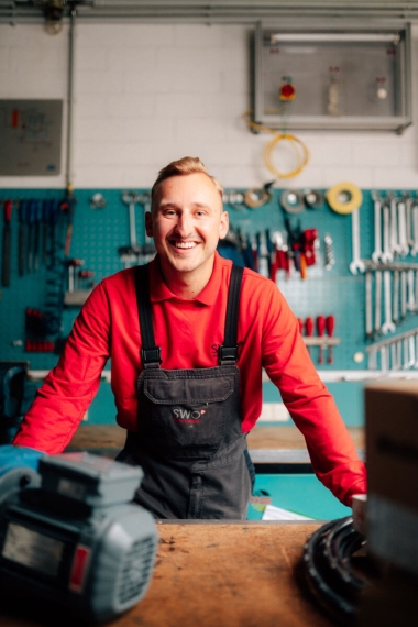 Ein breit lächelnder Handwerker in rotem Pullover und dunkler Latzhose steht in einer gut ausgestatteten Werkstatt hinter seiner Werkbank.