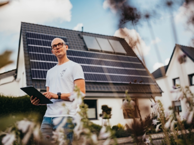 Ein Mann steht mit einem Tablet in der Hand vor einem Haus mit Photovoltaik-Anlage auf dem Dach