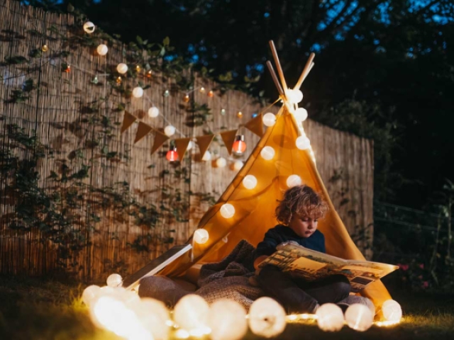 Ein Junge sitzt in einem Stoffzelt mit Lichterkette in einem Garten und liest ein Buch