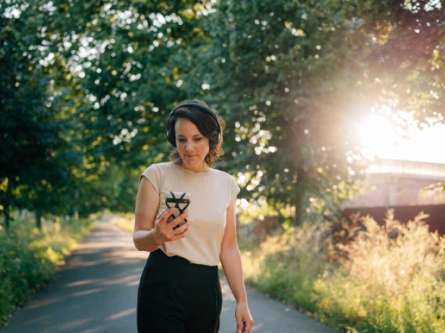 Eine Frau geht im Sonnenuntergang unter Bäumen spazieren mit Kopfhörern auf den Ohren und einem Handy in der Hand