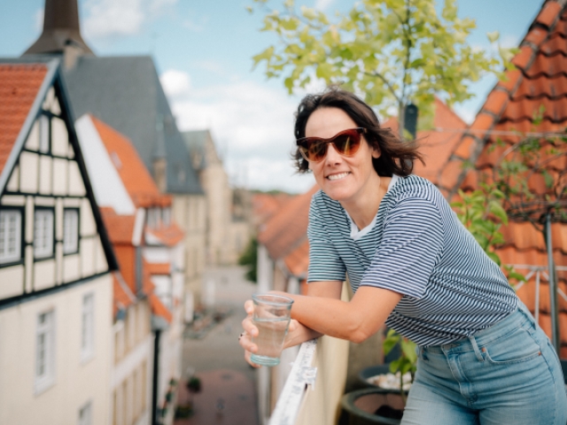 Eine Frau mit Sonnenbrille lehnt am Geländer einer Terrasse über den Dächern einer Stadt und hält ein Glas Wasser in der Hand 