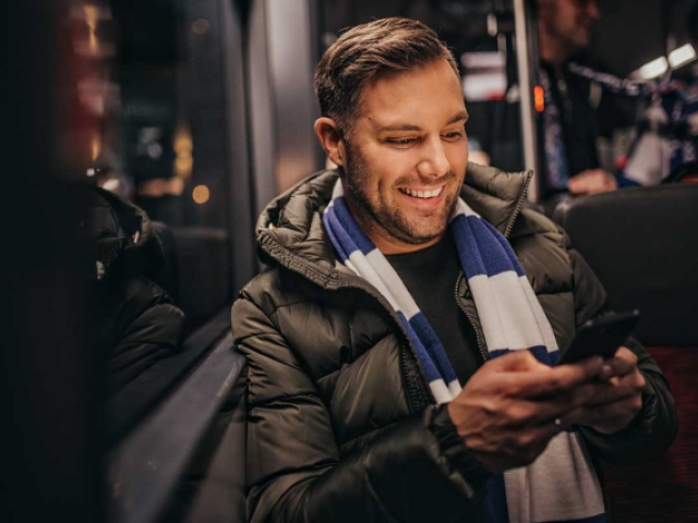 Ein Mann mit Fanschal des Vfl Osnabrück sitzt in einem Bus und schaut lächelnd auf ein Handy