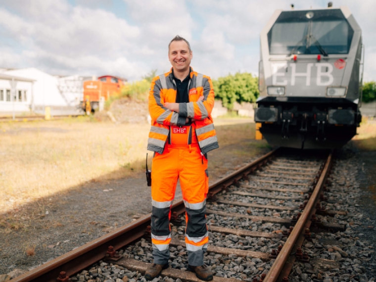 Ein lächelnder EHB-Mitarbeiter steht mit verschränkten Armen in kompletter orangefarbener Sicherheitskleidung auf den Gleisen vor einer großen grauen Lokomotive.